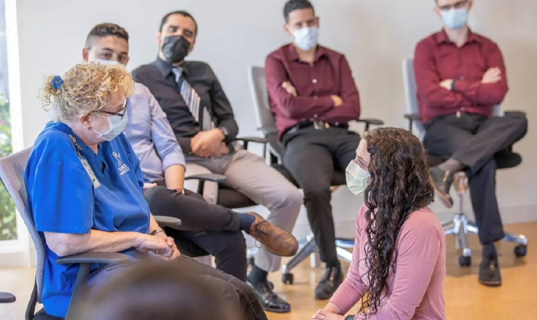 Group seated in semicircle wearing face coverings and watching interaction between two women  